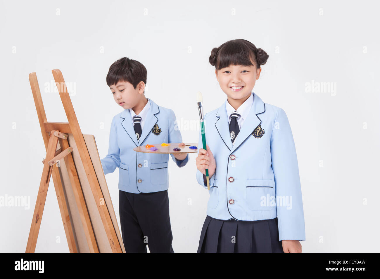 Grundschule Mädchen starrte nach vorne mit einem Lächeln hält einen Pinsel und junge zeichnen mit Staffelei hinter ihr sowohl in der Schule Stockfoto