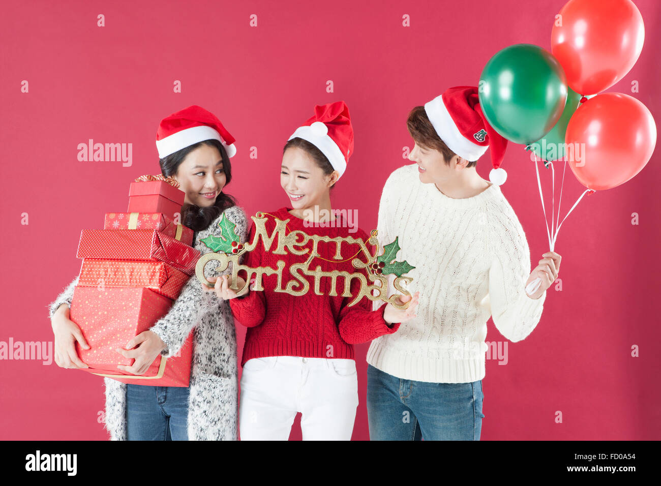 Drei junge Erwachsene in Nikolausmütze Abhaltung von Geschenk-Boxen, Frohe Weihnachten Zeichen und rote und grüne Luftballons auf rotem Grund Stockfoto