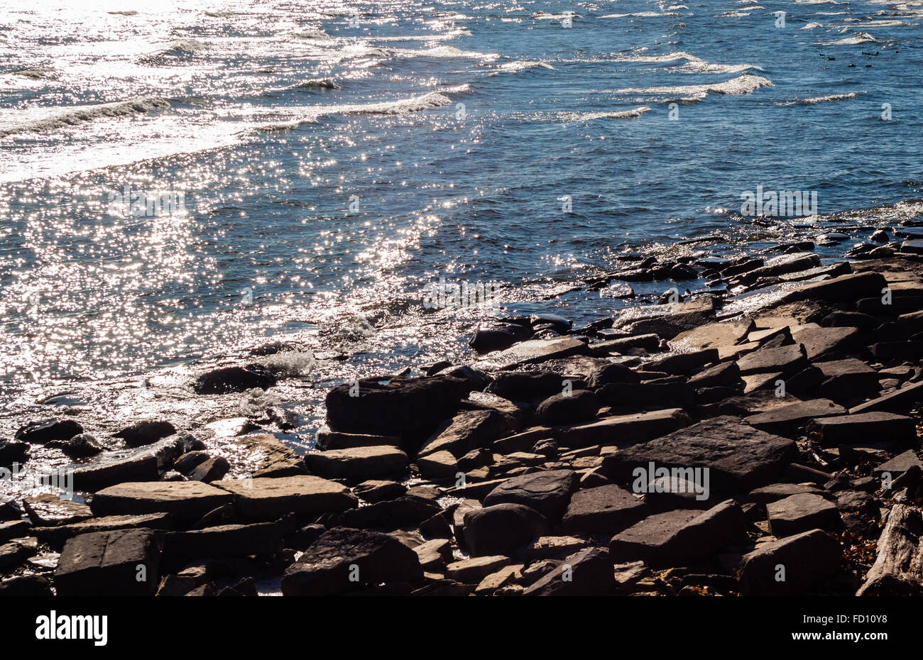 Braunen Felsen und Steine diagonal im Water's Edge, mit kleinen Wellen brechen und Wasser reflektiert Sonnenlicht. Stockfoto