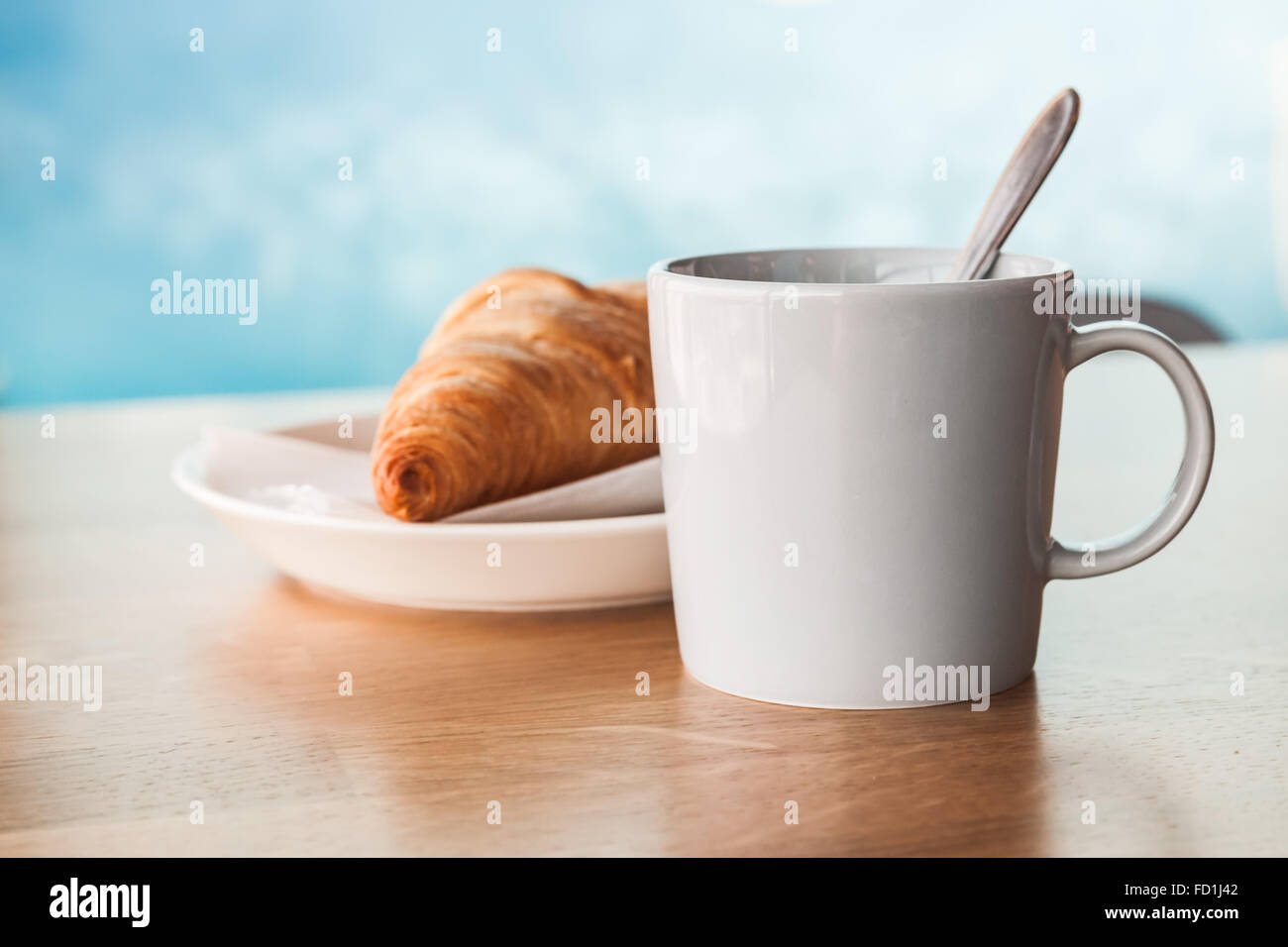 Cappuccino mit Croissant. Tasse Kaffee mit Milchschaum steht auf Holztisch in Cafeteria über blauen verschwommenen Hintergrund, closeu Stockfoto