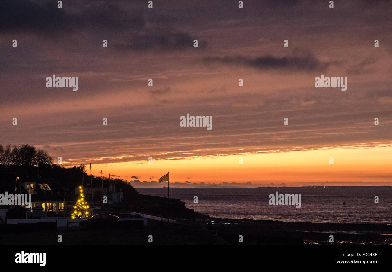 ein Streifen von leuchtend orange Licht quer durch eine Sonne set dunklen Himmel an der Küste mit einer außen Weihnachten Pinie bedeckt in Lichterketten Weihnachten Stockfoto