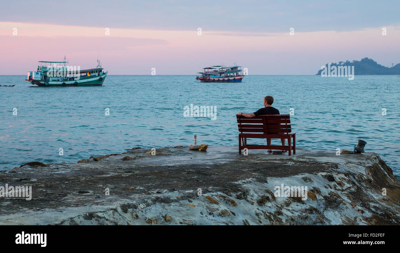 Einsamer Mann sitzt auf einer Bank an der Küste beobachten die Boote während des Sonnenuntergangs. Stockfoto