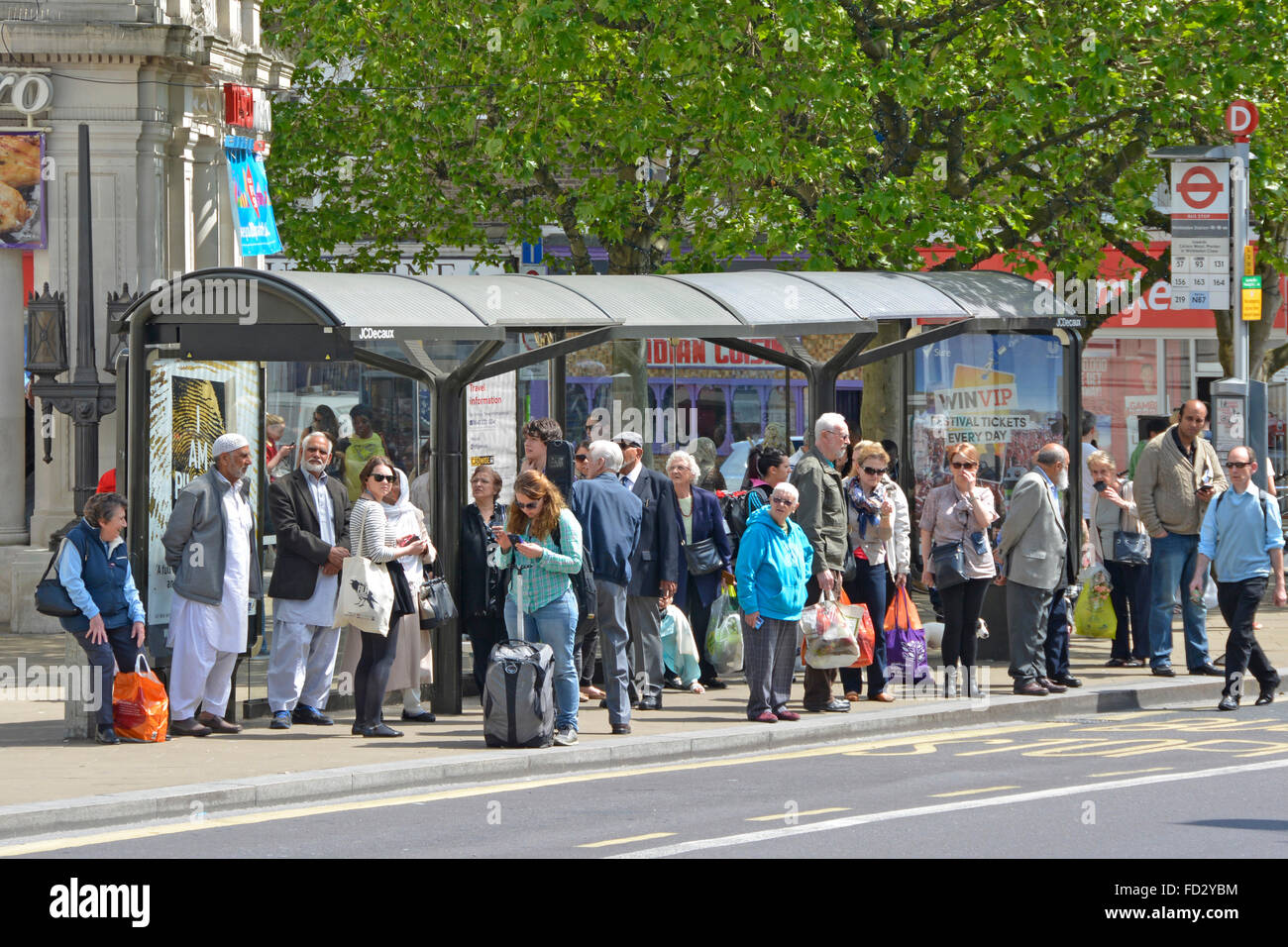 Gruppe von Menschen, einige mit Einkaufstaschen stehen & warten auf Transport for London geschäftige beliebte Bushaltestelle mit Schutz im Stadtzentrum Wimbledon UK Stockfoto