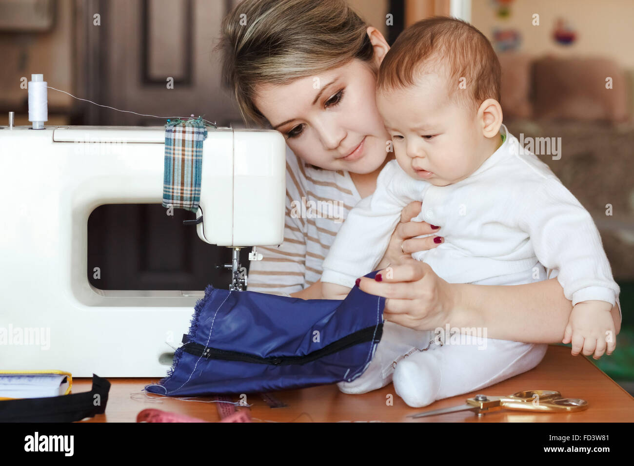 Baby mit seiner Mutter Näherin, Heimat, natürliches Licht, Kleinkind, die neugierig auf die Nähmaschine. Kinderbetreuung und Arbeit zu Hause. Stockfoto
