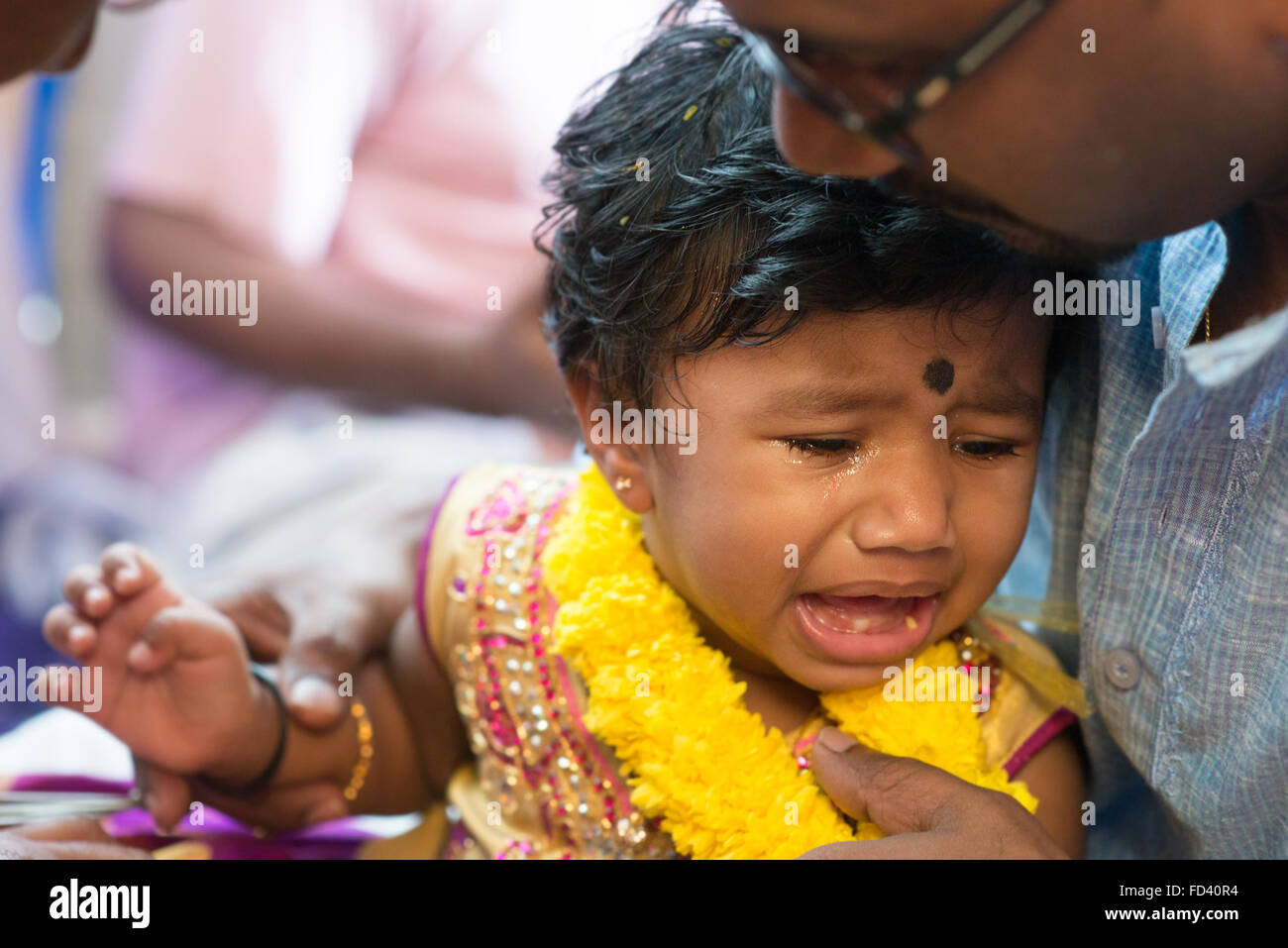Vater verwöhnt seine Babymädchen, weinend in die Karnavedha-Ereignisse. Traditionelle indische Hindus Ohrlochstechen Zeremonie. Indien-speci Stockfoto