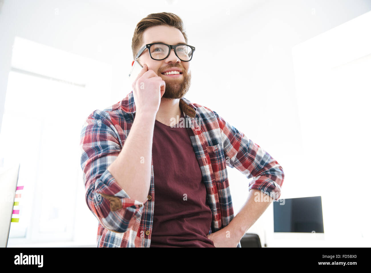 Fröhliche junge bärtiger Mann in Gläsern telefonieren mit Handy im Büro und lächelnd Stockfoto