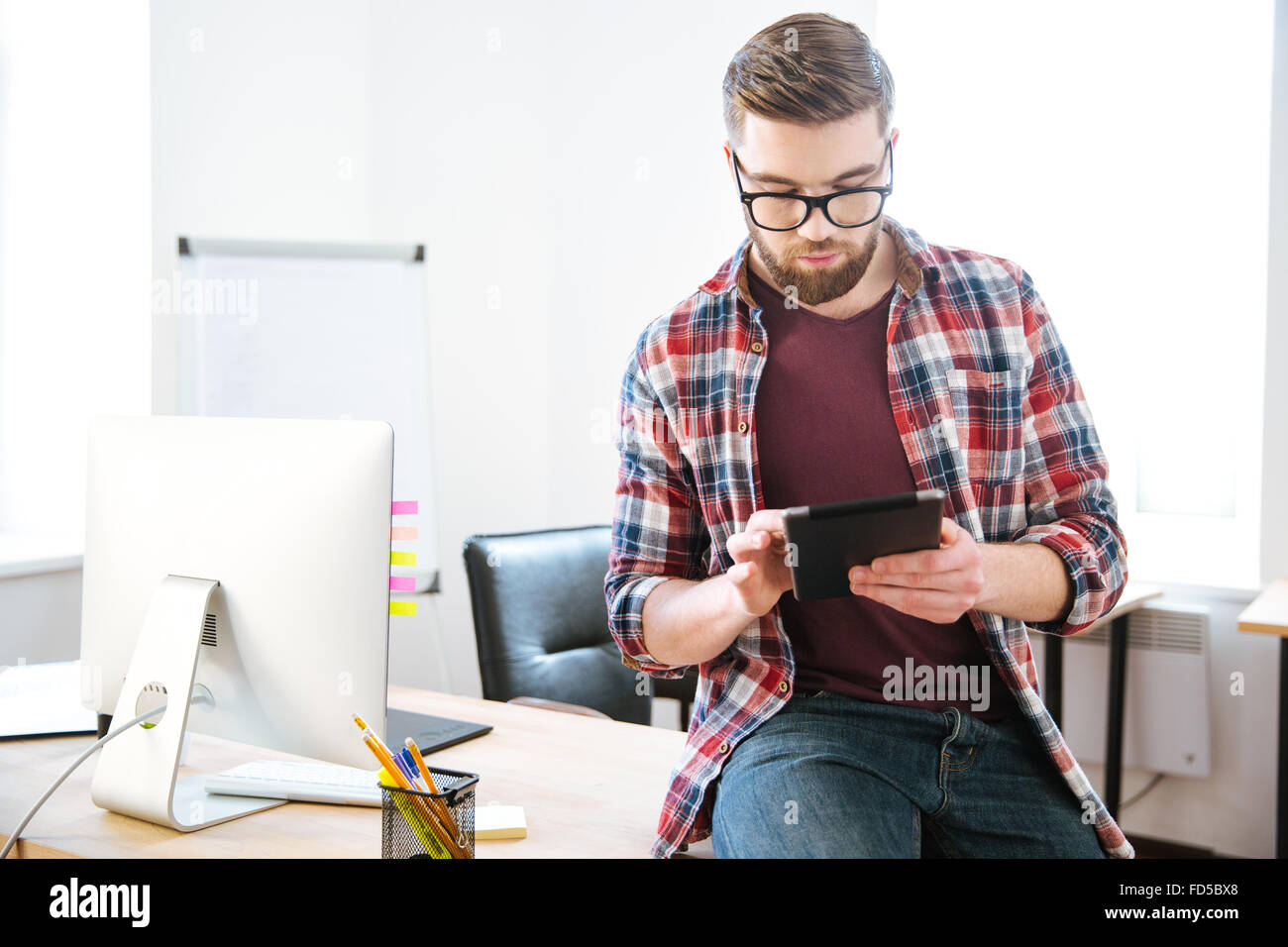 Nachdenklichen jungen Mann mit Bart konzentriert in Gläser auf dem Tisch im Büro sitzen und mit tablet Stockfoto