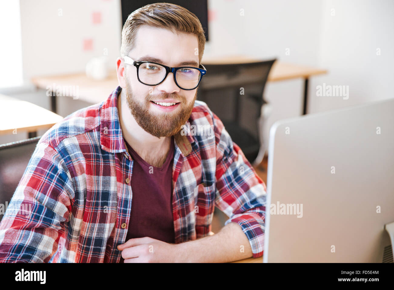 Glücklichen hübscher junger Mann mit Perle im karierten Hemd und Brille sitzen am Arbeitsplatz und die Arbeit mit computer Stockfoto