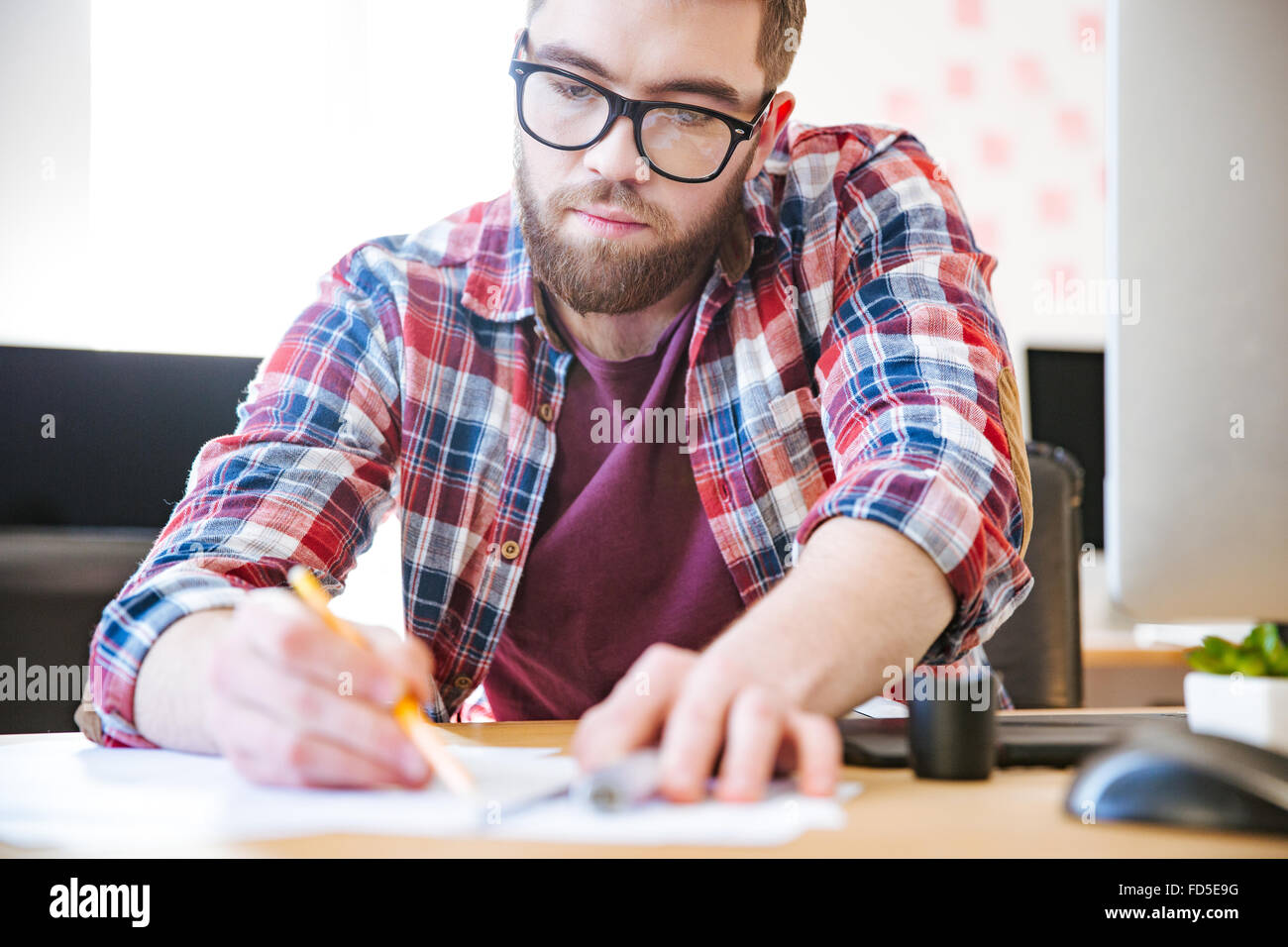 Gut aussehend ernster Mann im karierten Hemd und Brille sitzen und schreiben im Büro Stockfoto