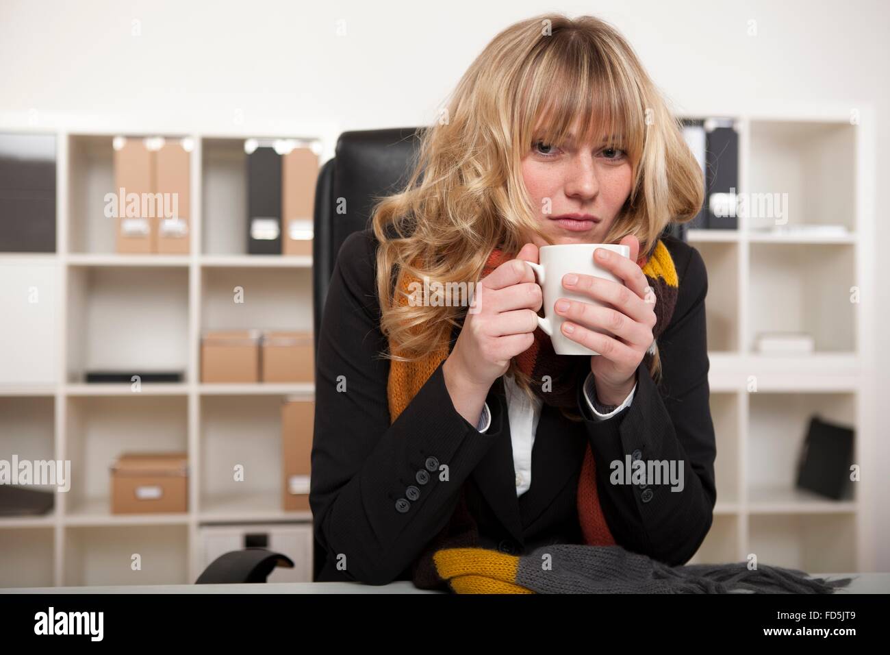 Kalten Geschäftsfrau mit einer Tasse heißen Kaffee aufwärmen, als sie an ihrem Schreibtisch im Büro trägt einen Schal gestrickt Winter sitzt. Stockfoto