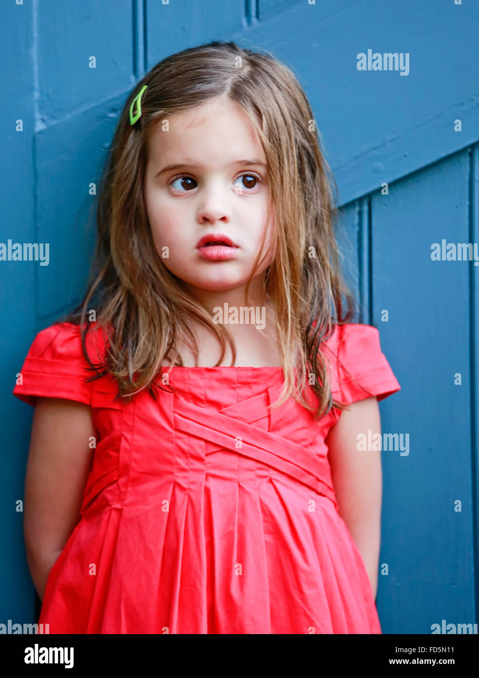 Kleines Mädchen in einem hellen, roten Kleid gegen ein blaues Tor mit einem ängstlich Blick auf ihrem Gesicht. Stockfoto