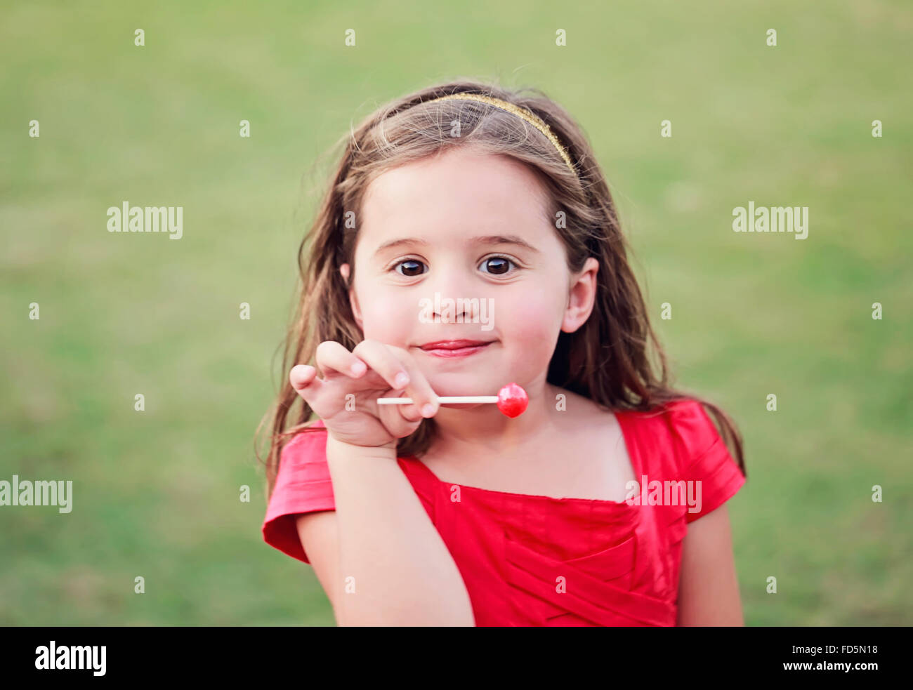 Kleines Mädchen in einem leuchtend roten Kleid einen leuchtenden roten Lutscher während lächelnd in die Kamera halten. Stockfoto