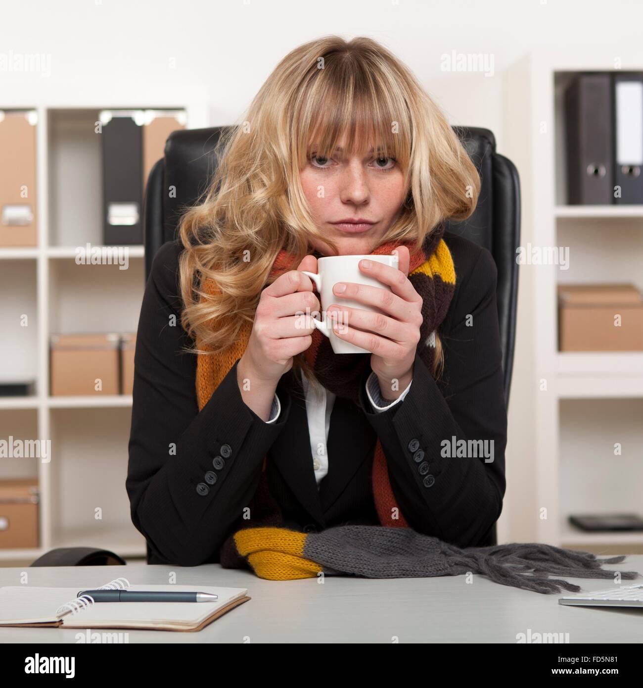 Junge Mädchen bei der Arbeit sitzt an ihrem Schreibtisch im Büro mit einem warmen Winter Schal um den Hals, genießen Sie eine Tasse heißen Kaffee Stockfoto