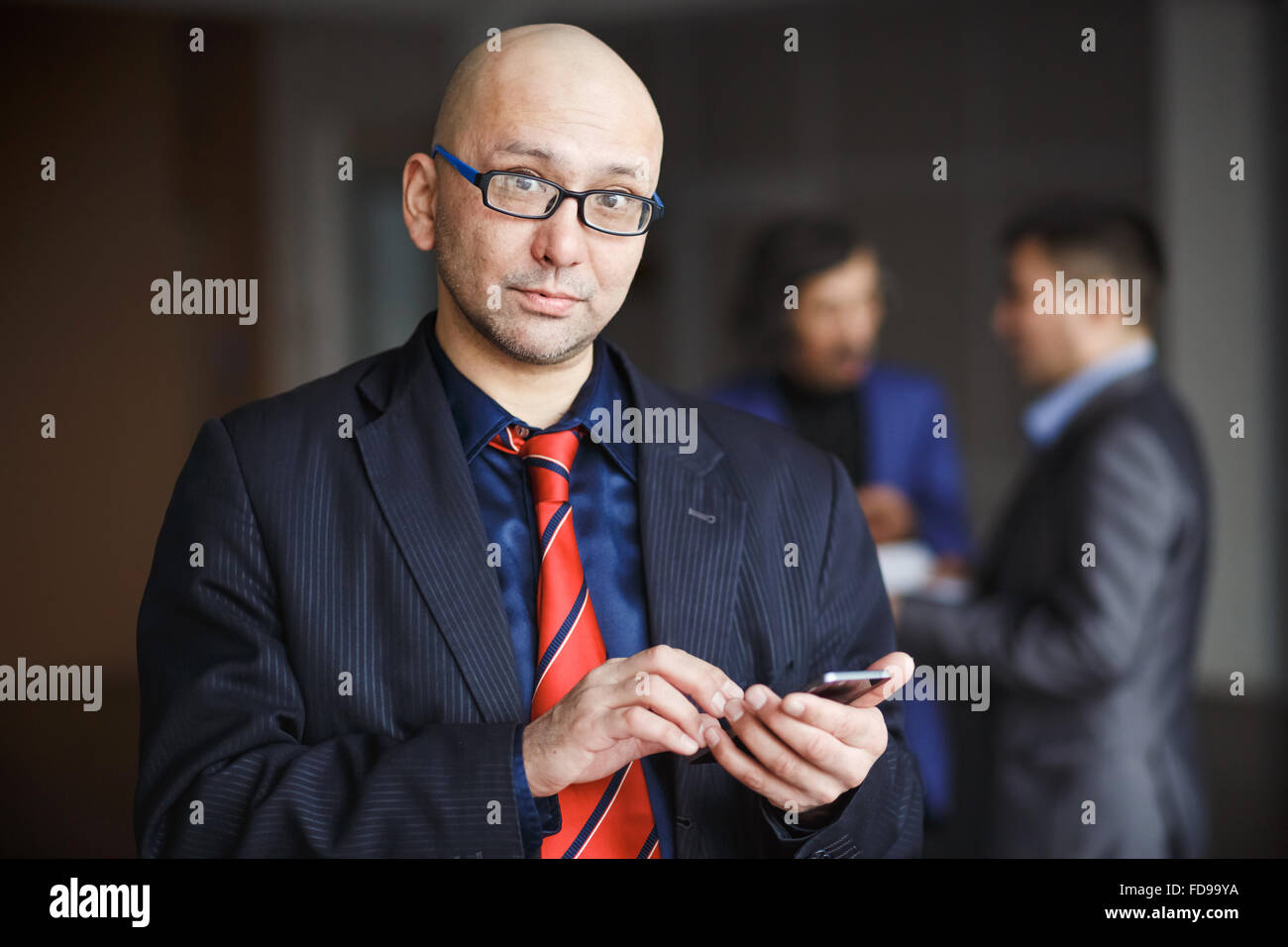 Porträt des Mannes mit Brille Glatze Geschäftsmann mit Handy in der hand gekleidet gestreiften Anzug und roter Krawatte, steht drinnen Bürogebäude. Er schaut direkt in die Kamera. Bei Hintergrund Man diskutieren. Stockfoto