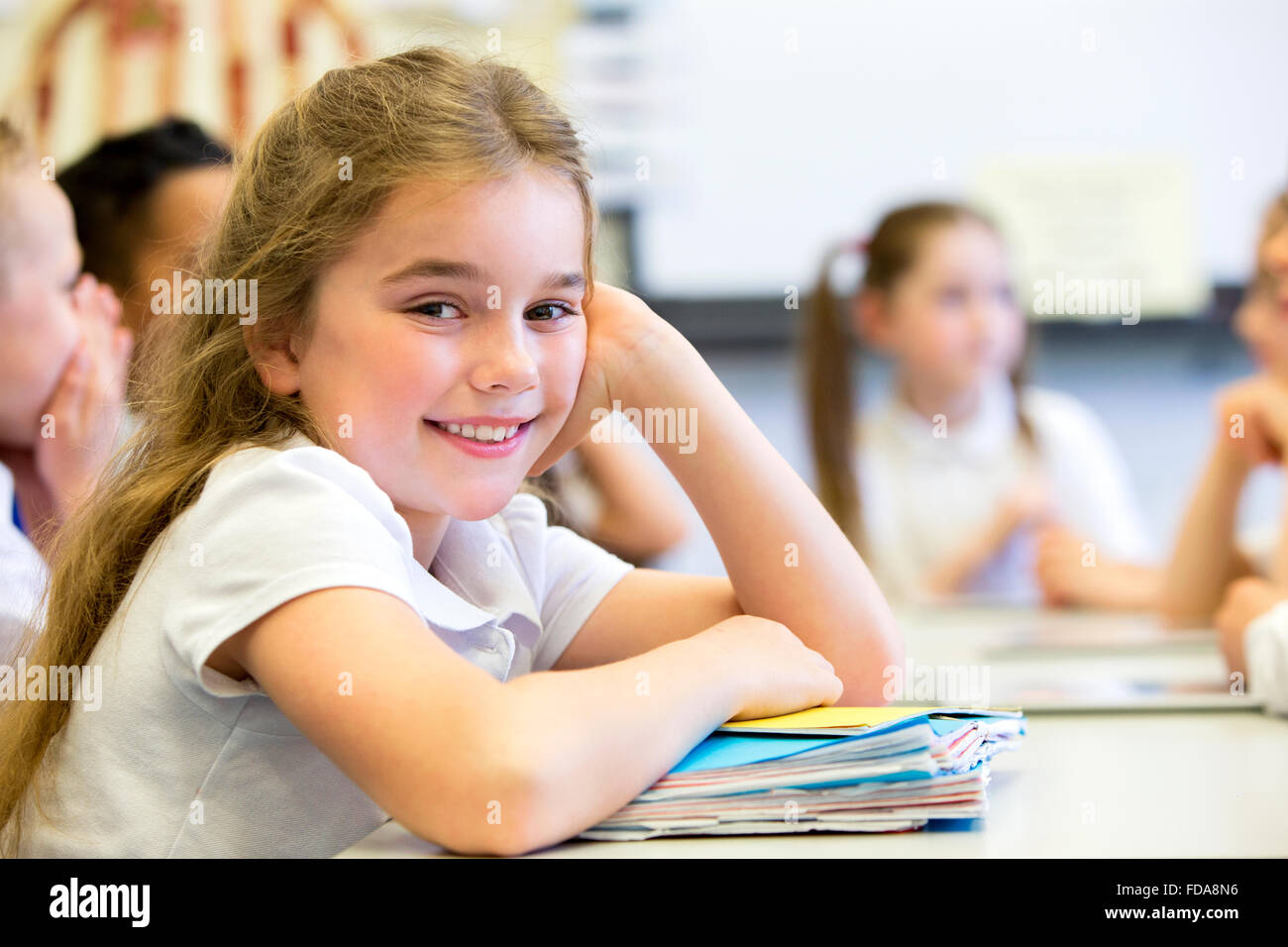 Schulmädchen lächelt in die Kamera, als sie an ihrem Schreibtisch beim stillstehen auf einen großen Haufen Papierkram sitzt. Stockfoto