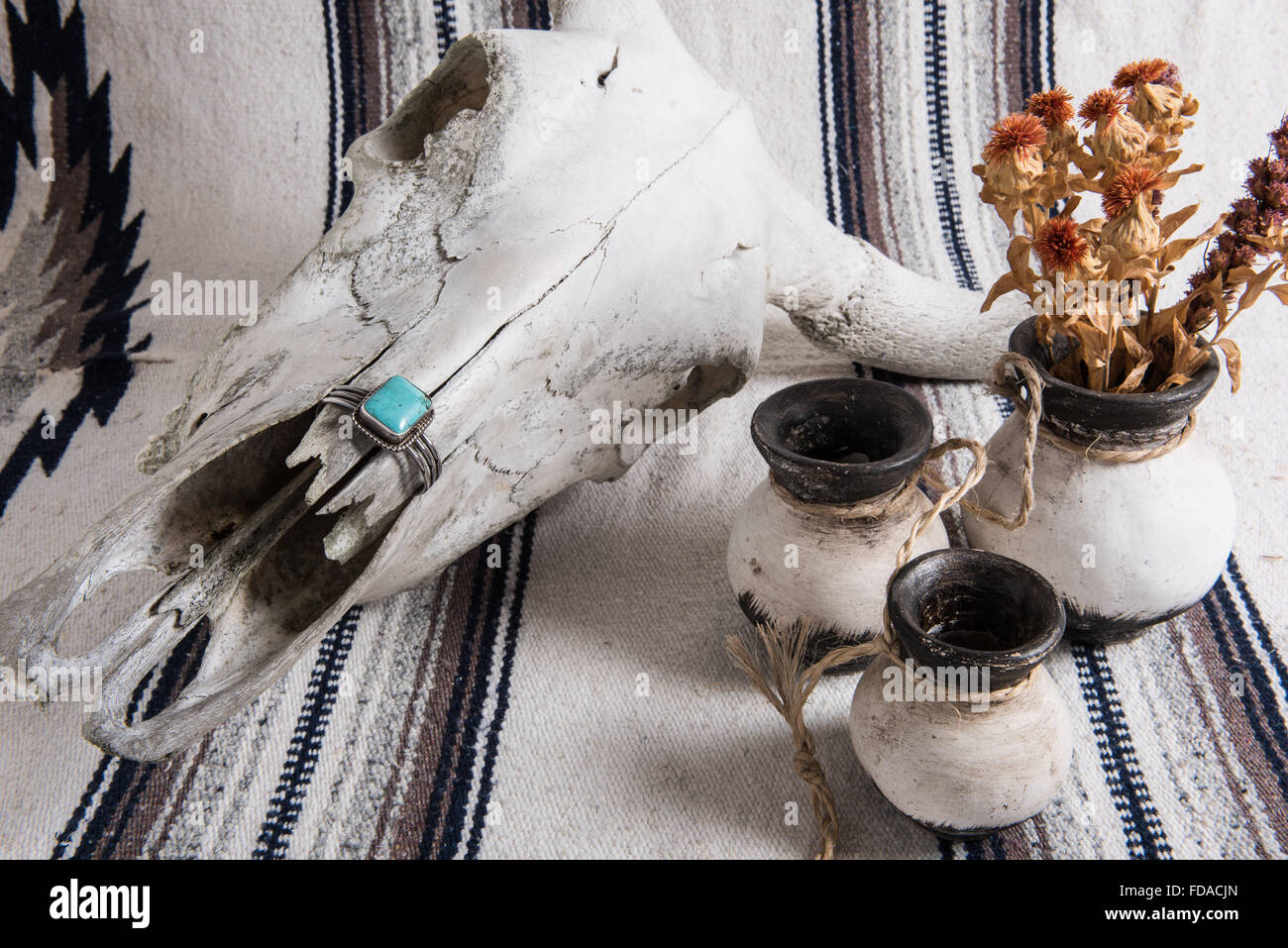 Verwitterter Kuhschädel, türkisfarbenes und silbernes Manschettenarmband, schwarz-weiß bemalte mexikanische Keramik mit getrockneten Blumen auf einer mexikanischen gestreiften Decke. Stockfoto