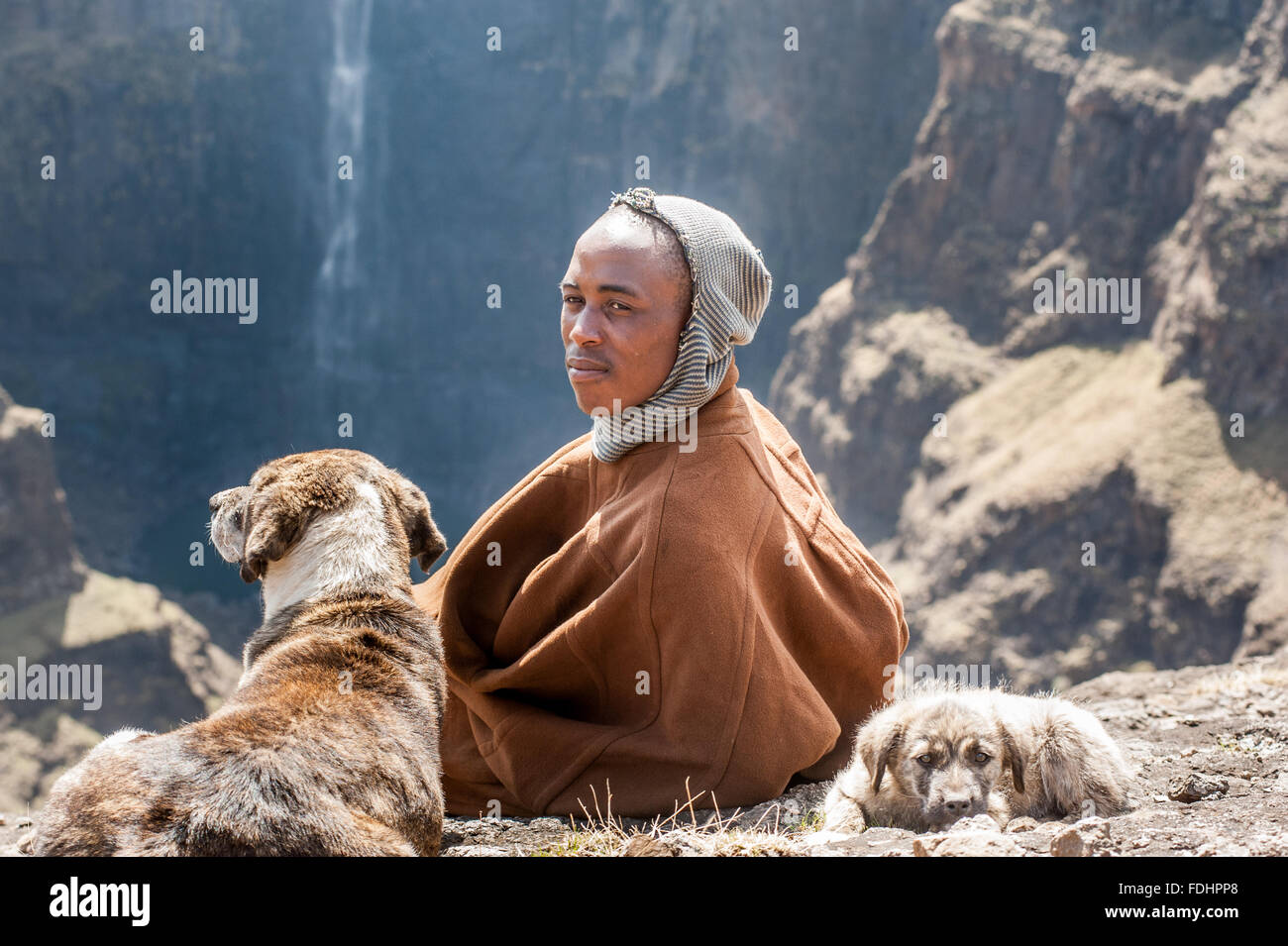 Ein Hirte, eingewickelt in eine Decke sitzt mit seinen Hunden gegenüber Maletsunyane-Wasserfall in Somenkong, Lesotho, Afrika Stockfoto