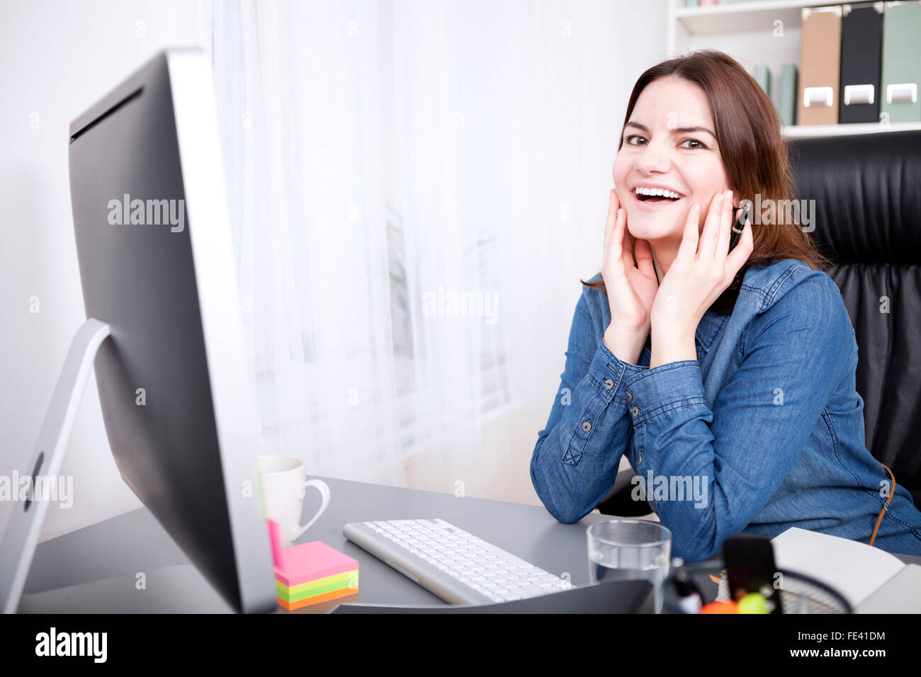 Happy Büro Frau gelehnt auf dem Tisch in ihrem Büro mit Händen im Gesicht und auf der Suche in die Kamera. Stockfoto