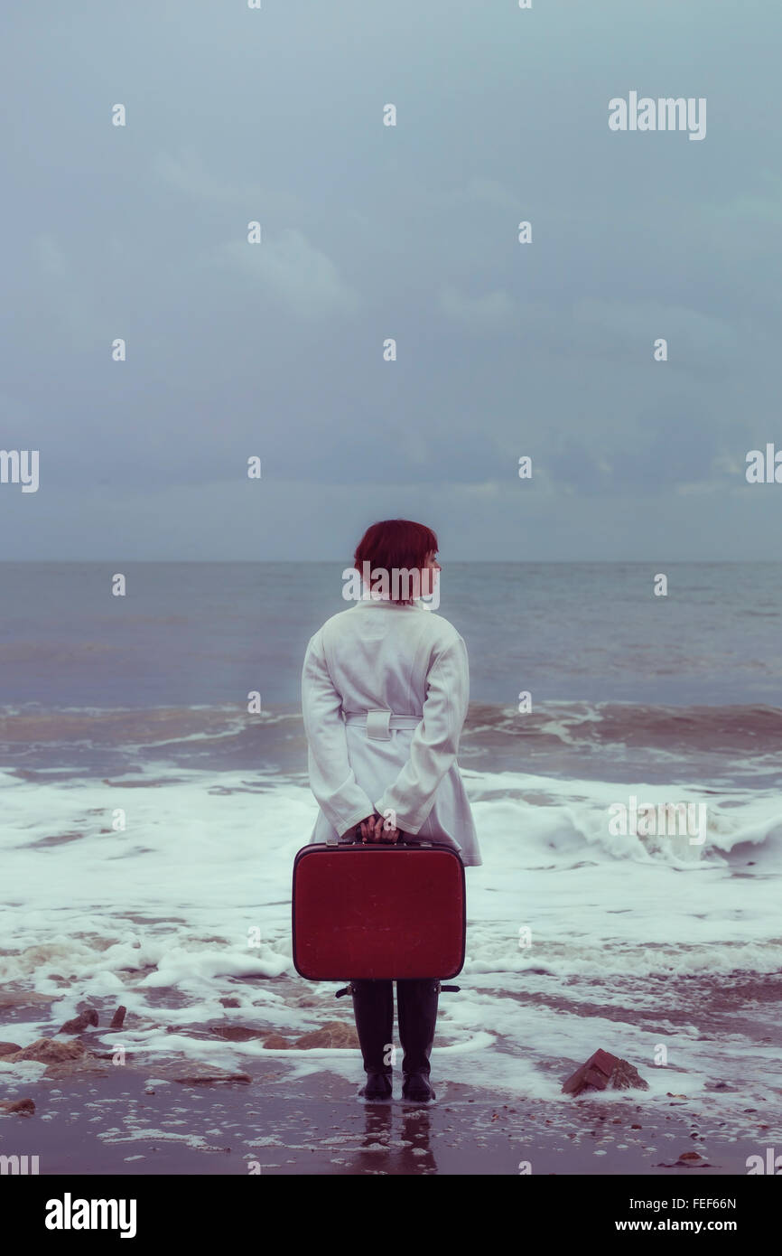 eine Frau in einem weißen Mantel mit einem roten Koffer am Meer im winter Stockfoto