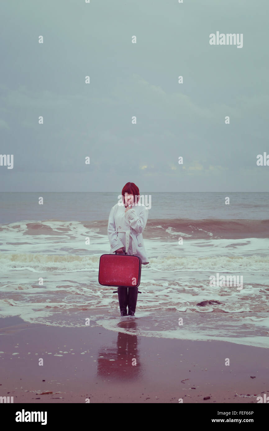 eine Frau in einem weißen Mantel mit einem roten Koffer am Meer im winter Stockfoto