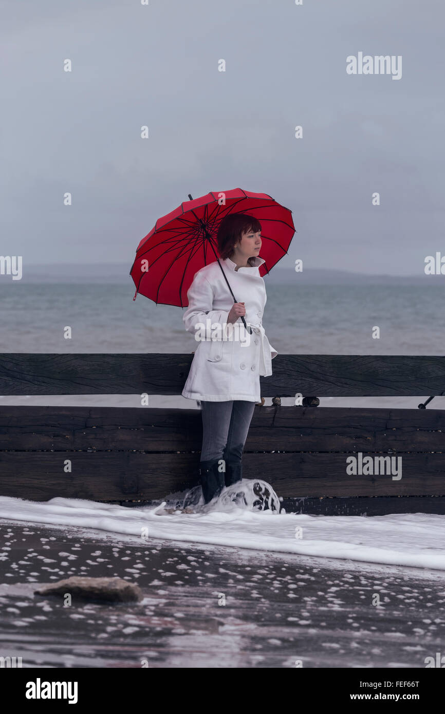 eine Frau in einem weißen Mantel mit einem roten Regenschirm am Meer im winter Stockfoto