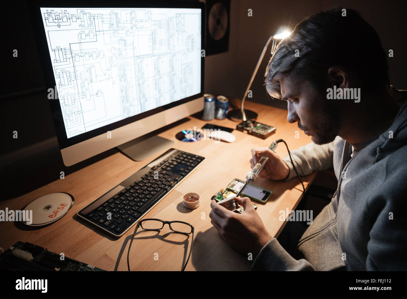 Ernst gut aussehenden jungen Mann mit Lötkolben zur Befestigung Smartphone sitzen im dunklen Raum Stockfoto