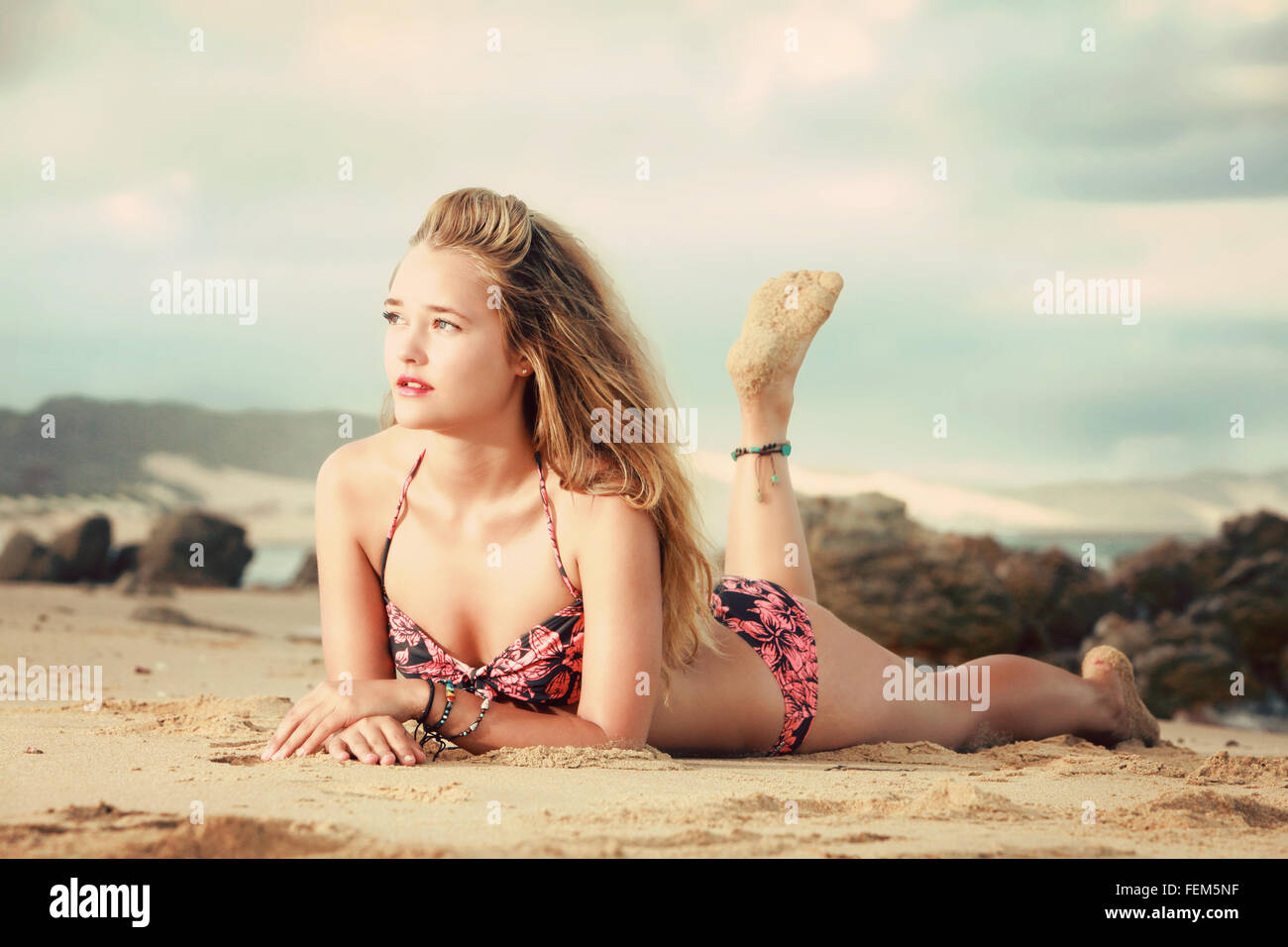 Eine schöne Blondine liegt auf dem Bauch auf einem Felsen am Strand in Afrika. Sie hat einen Bikini und weißen Shorts am Strand Stockfoto
