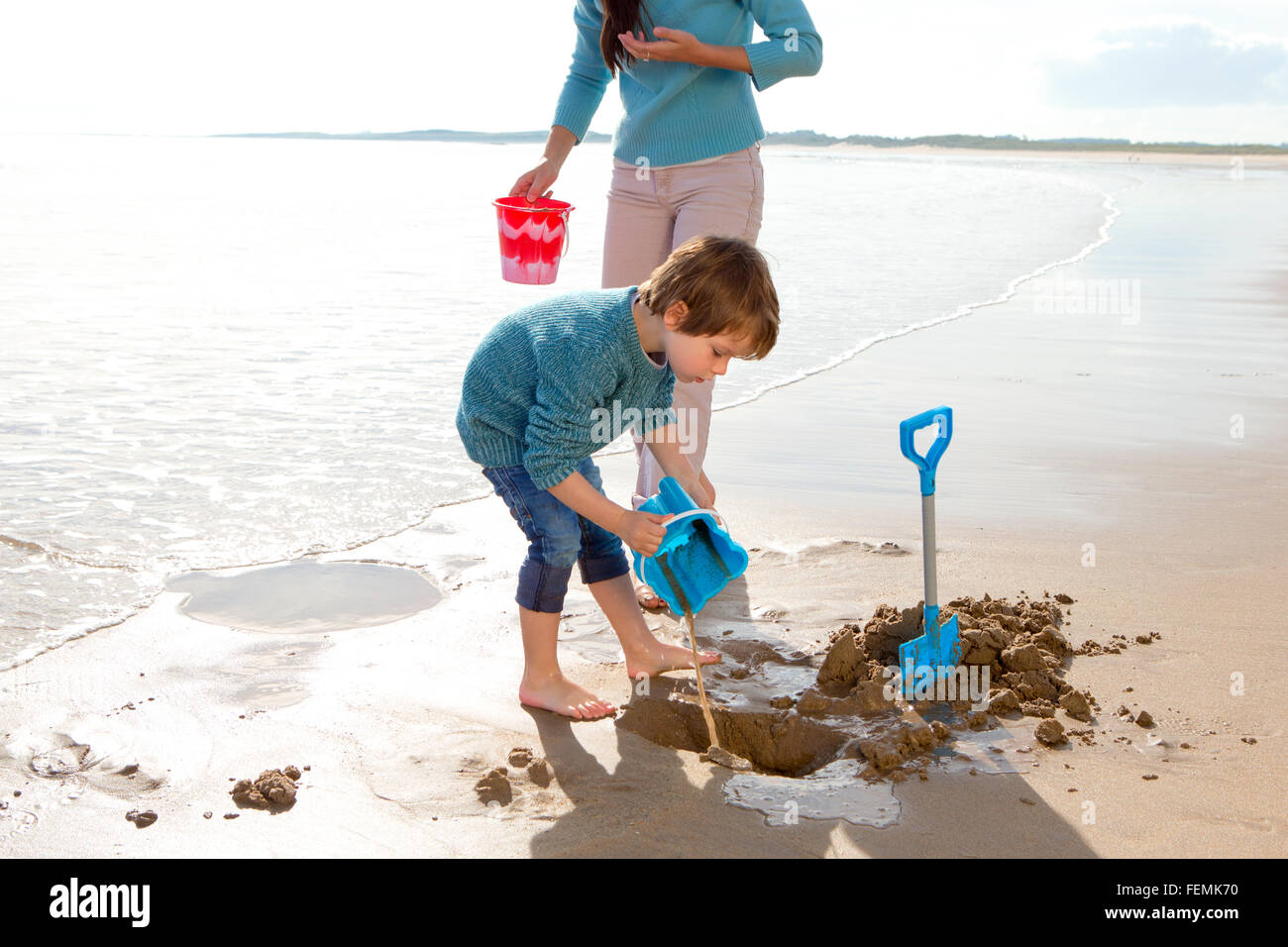 Kleiner Junge spielt am Strand mit einem Eimer und Schaufel mit seiner Mutter. Stockfoto