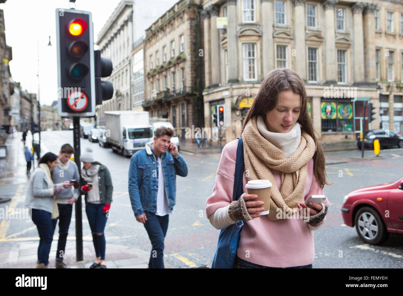 Eine junge Frau kann zu Fuß entlang einer Stadtstraße mit einem Smartphone gesehen werden. Andere junge Erwachsene sind im Hintergrund. Stockfoto