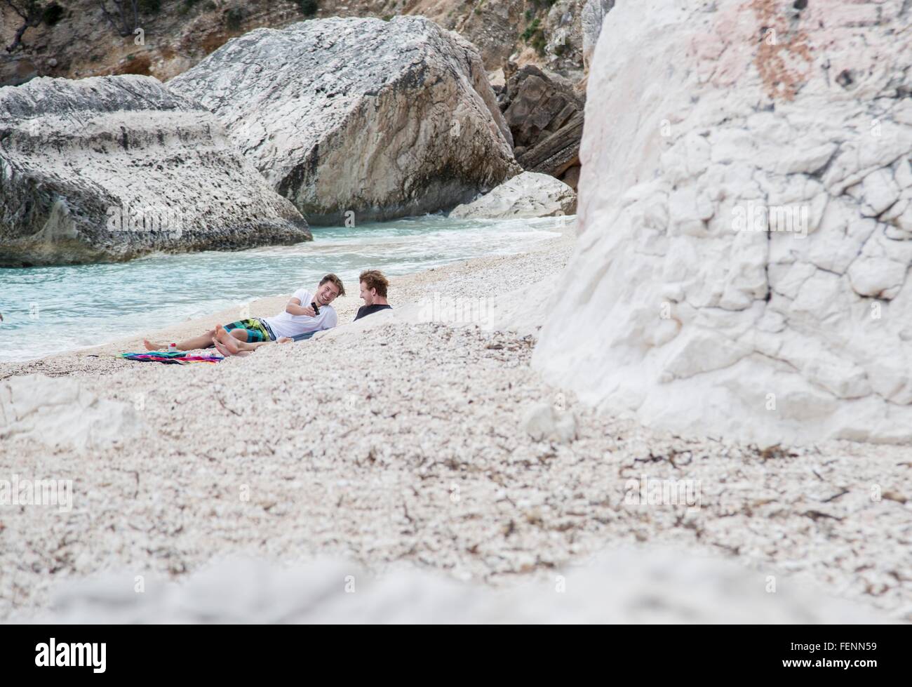 Junge Männer entspannend auf felsigen Strand mit Smartphone, Golfo di Orosei, Sardinien, Italien Stockfoto