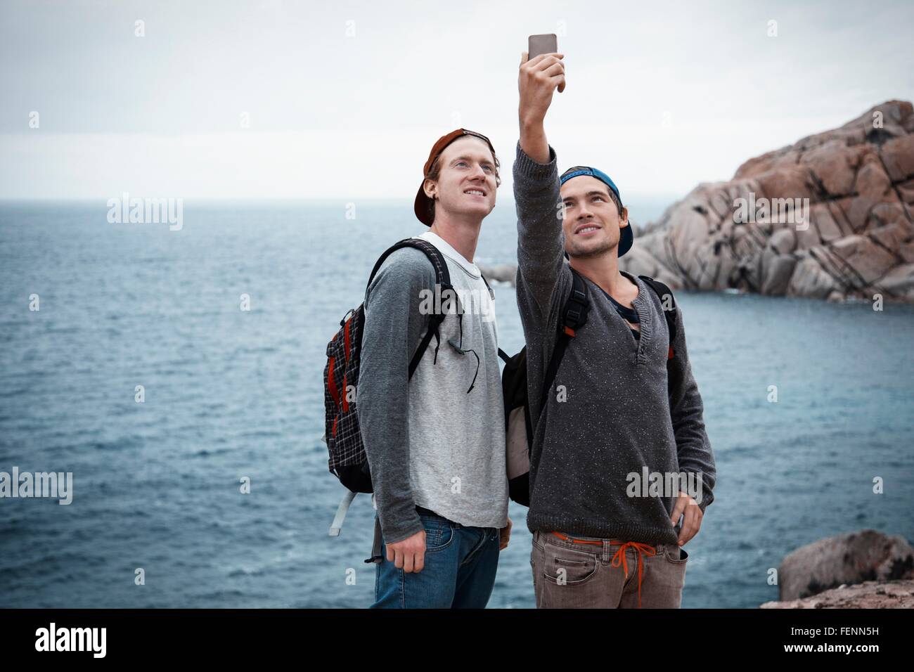 Junge Männer vor Ozean mit Smartphone zu Selfie lächelnd, Costa Smeralda, Sardinien, Italien Stockfoto