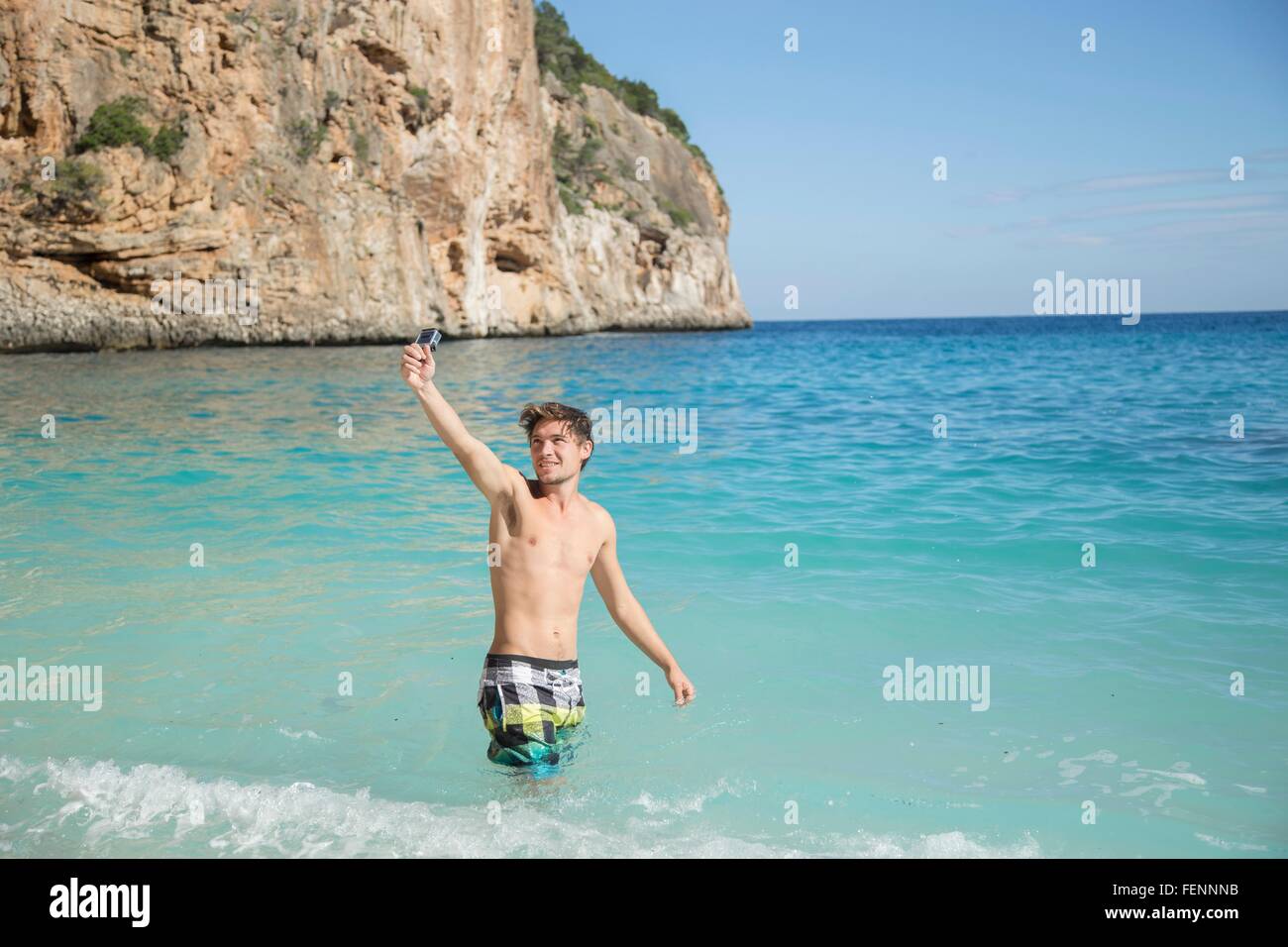 Junger Mann im Ozean erhobenem Arm mit Kamera Lächeln, Golfo di Orosei, Sardinien, Italien Stockfoto