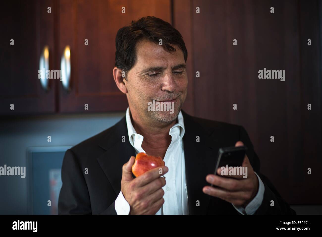 Reifer Mann mit Anzugjacke hält Apfel, lächelnd Smartphone betrachten Stockfoto