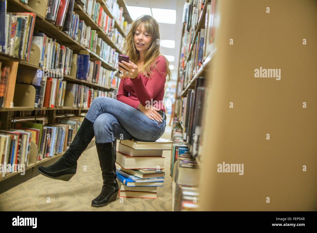 Reife Frau sitzen auf Stapel von Büchern in der Bibliothek lesen Smartphone Texte Stockfoto