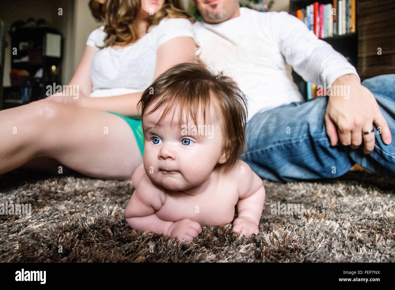 Mutter und Vater sitzen Rücken an Rücken auf Teppich mit Baby Girl auf Teppich wegsehen liegend Stockfoto