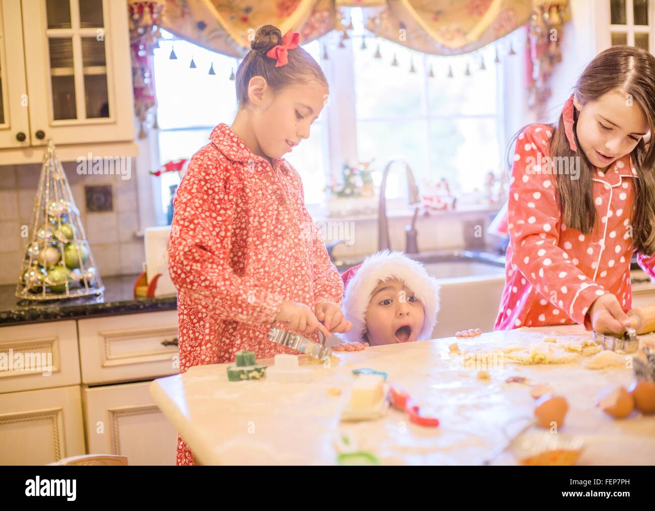 Junge in Santa Hut machen Cookies mit Schwestern peeking über Küchentheke Küche öffnen Mund Stockfoto