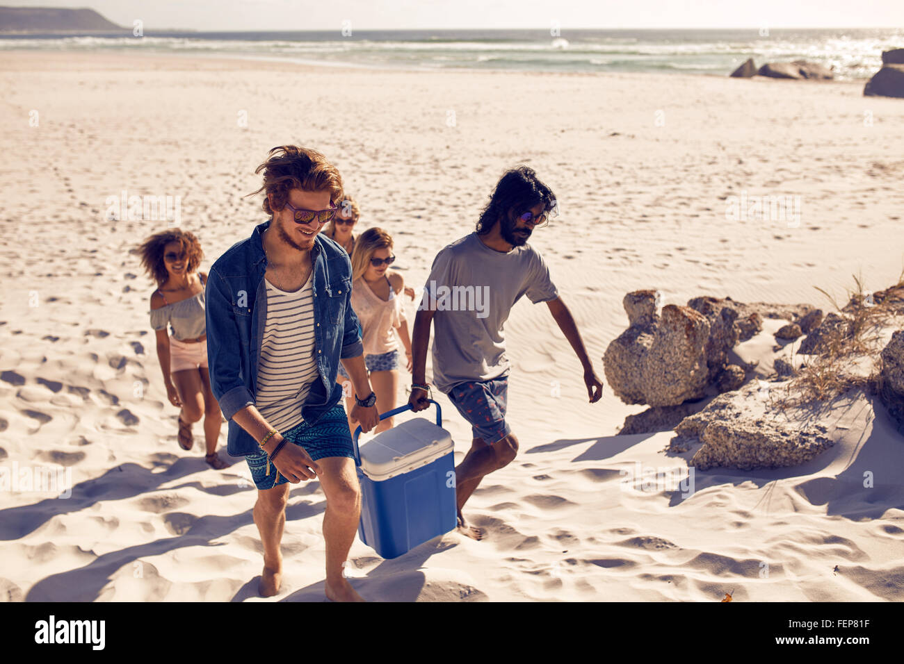 Gruppe von Menschen, die die Kühler, um am Strand zu feiern. Zwei junge Männer tragen Kühler mit Freundinnen auf der Rückseite, auf der Suche nach einem Stockfoto