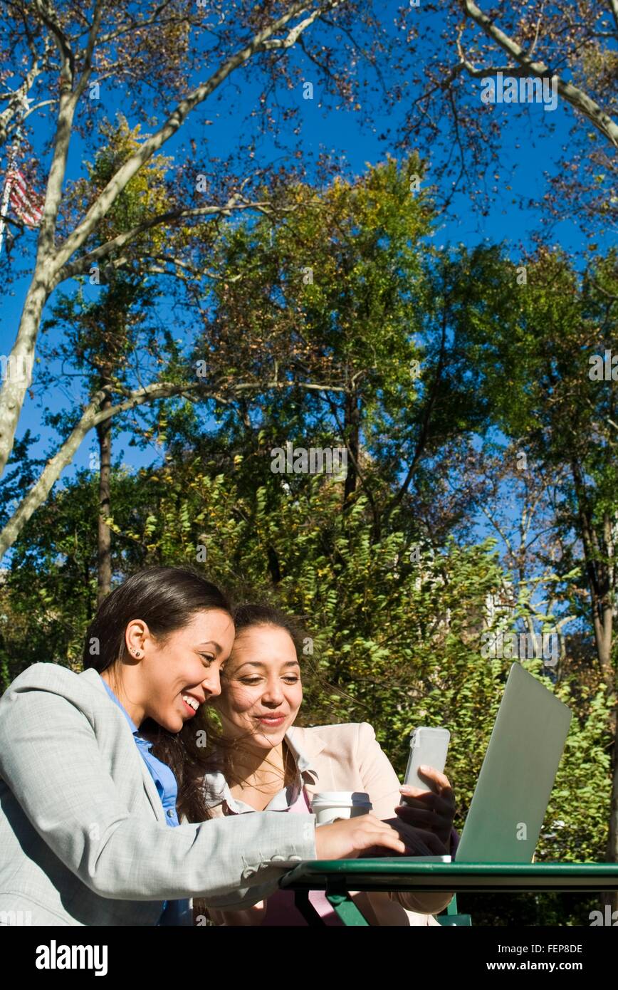 Junge Erwachsene weibliche Zwillinge mit Laptop und Smartphone im Stadtpark Stockfoto