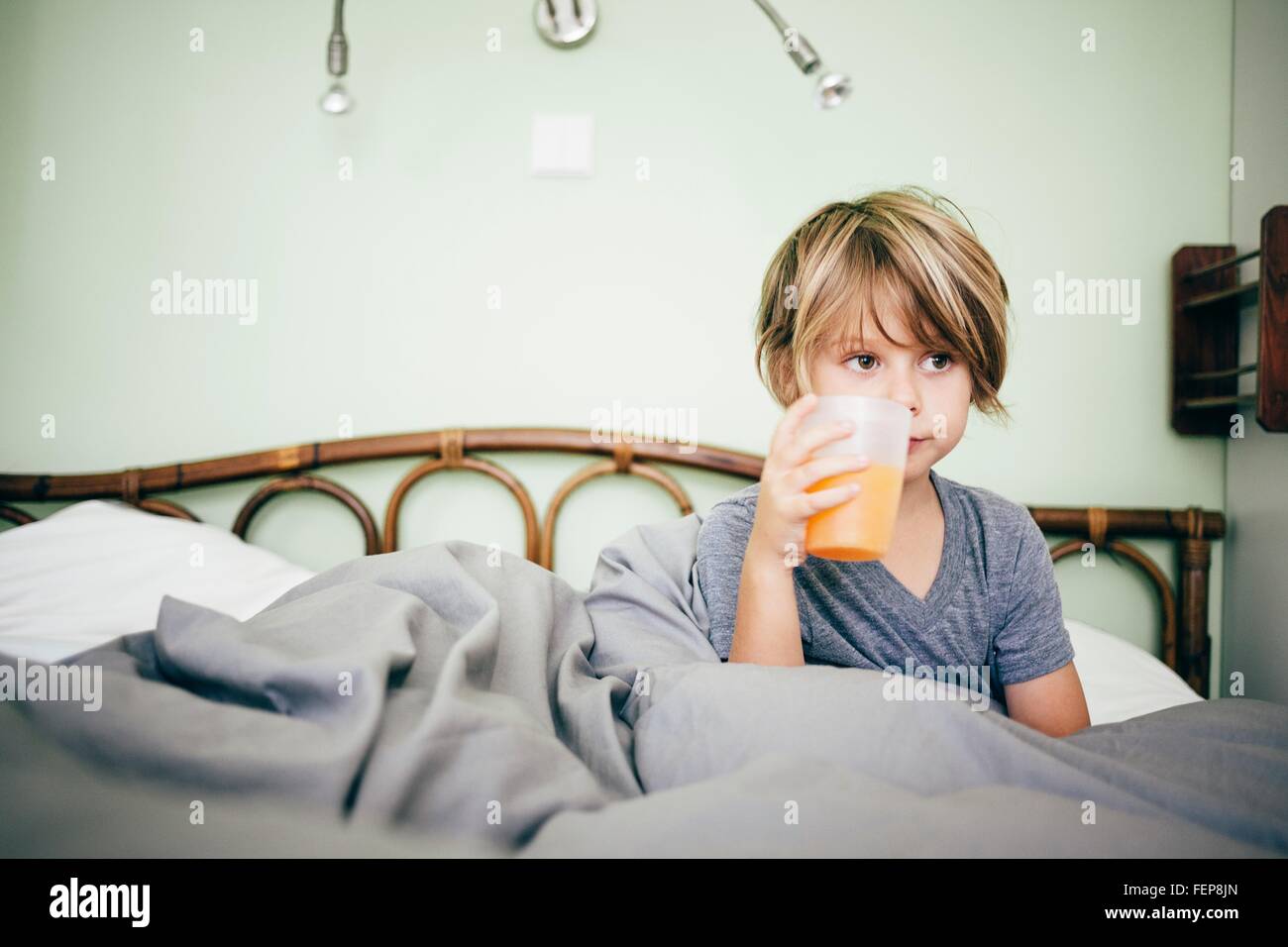 Junge sitzt im Bett trinken Becher Orangensaft, wegsehen, Bludenz, Vorarlberg, Österreich Stockfoto