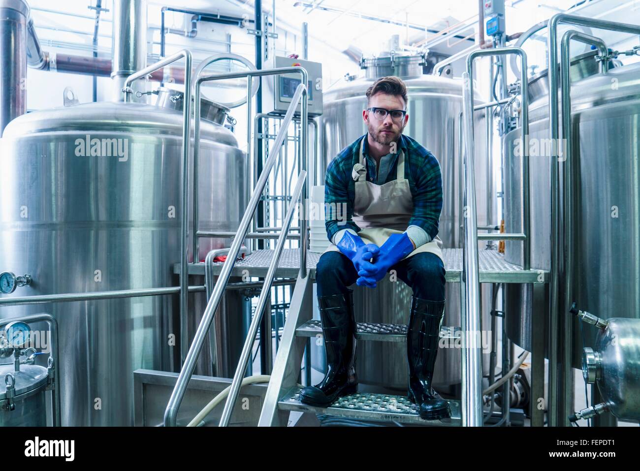 Junger Mann in der Brauerei tragen, Schürze, Handschuhe und Gummistiefel sitzen auf Stufen Blick in die Kamera Stockfoto