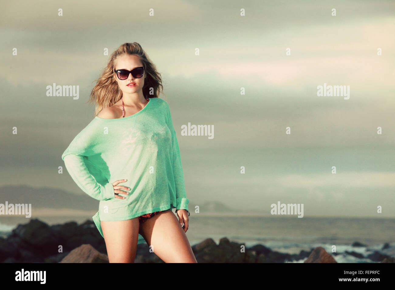 Eine attraktive junge blonde Dame, die mit einem grünen Trikot steht auf einem Felsen am Strand, wo die Flut in gekommen ist. Stockfoto