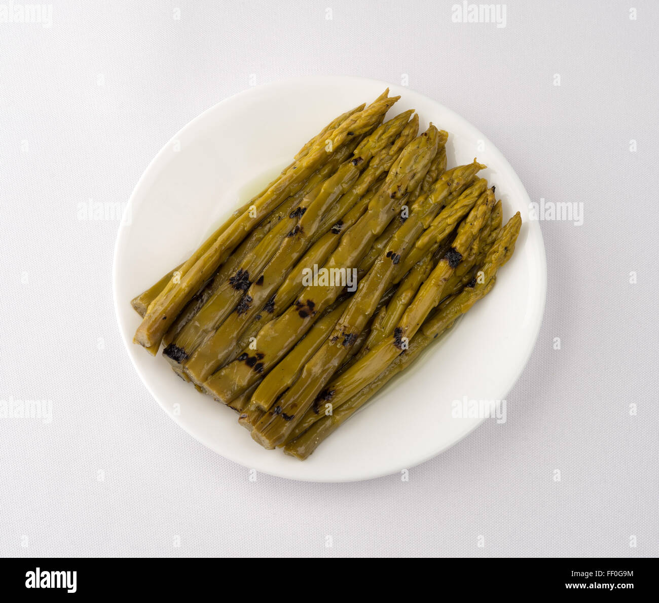 Eine Portion gegrillter Spargel getränkt in Sonnenblumenöl auf einem weißen Teller oben auf einem off White Tischdecke Stockfoto