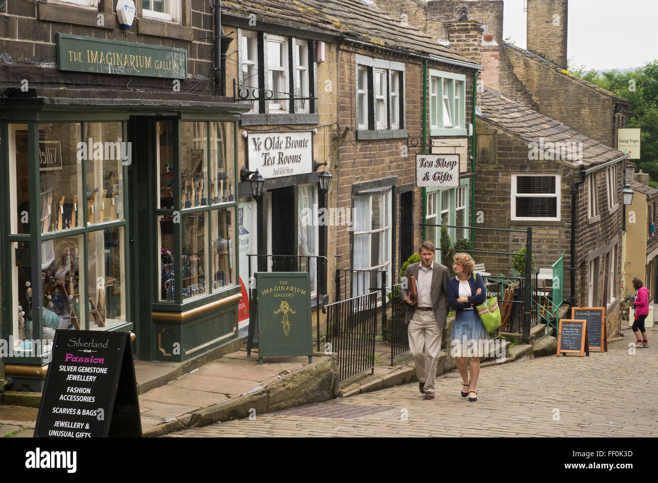 Paar zu Fuß bis steile Main Street, Haworth, West Yorkshire, England - malerischen, gepflasterten, gesäumt mit Geschäften, beliebt bei Bronte Land Besucher! Stockfoto