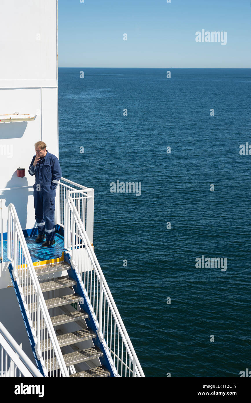 Seaman Maschinist in blauer Arbeitskleidung insgesamt Rauchen auf Treppe während der Zigarettenpause bei Sonnenschein mit der Ostsee Stockfoto