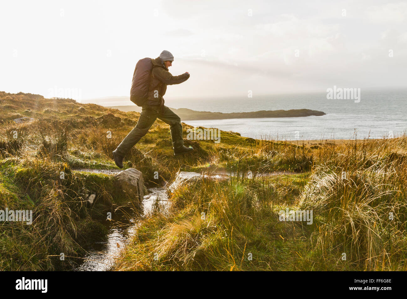 Ein Mann mit einem Rucksack und Winter Kleidung springen über einen kleinen Bach in eine exponierte Offenland. Stockfoto