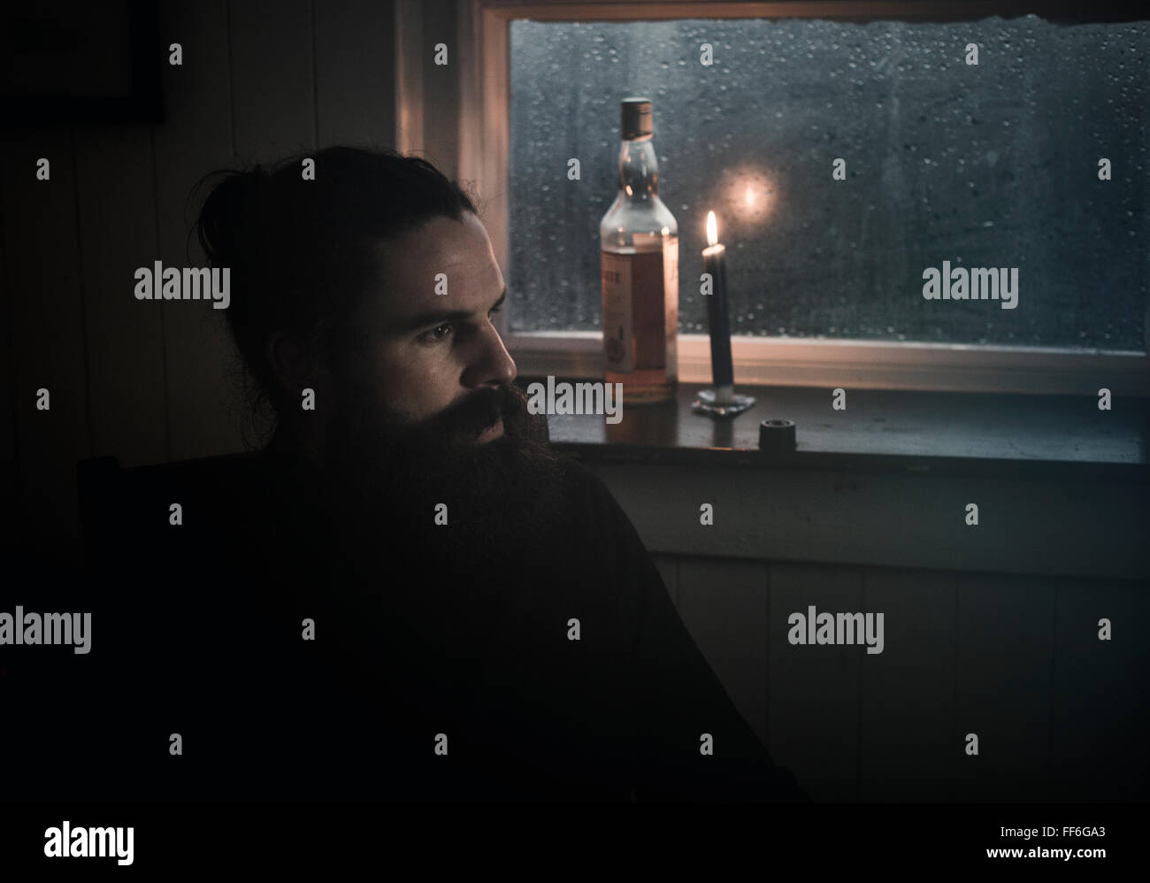 Ein Mann sitzt in der Dunkelheit durch ein Fenster im Kerzenlicht trinken aus einem kleinen Glas.  Eine Flasche neben ihm. Stockfoto