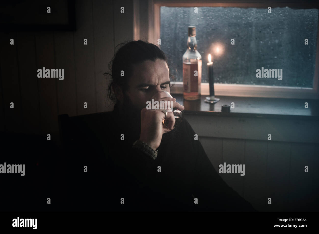 Ein Mann sitzt in der Dunkelheit durch ein Fenster im Kerzenlicht trinken aus einem kleinen Glas.  Eine Flasche neben ihm. Stockfoto