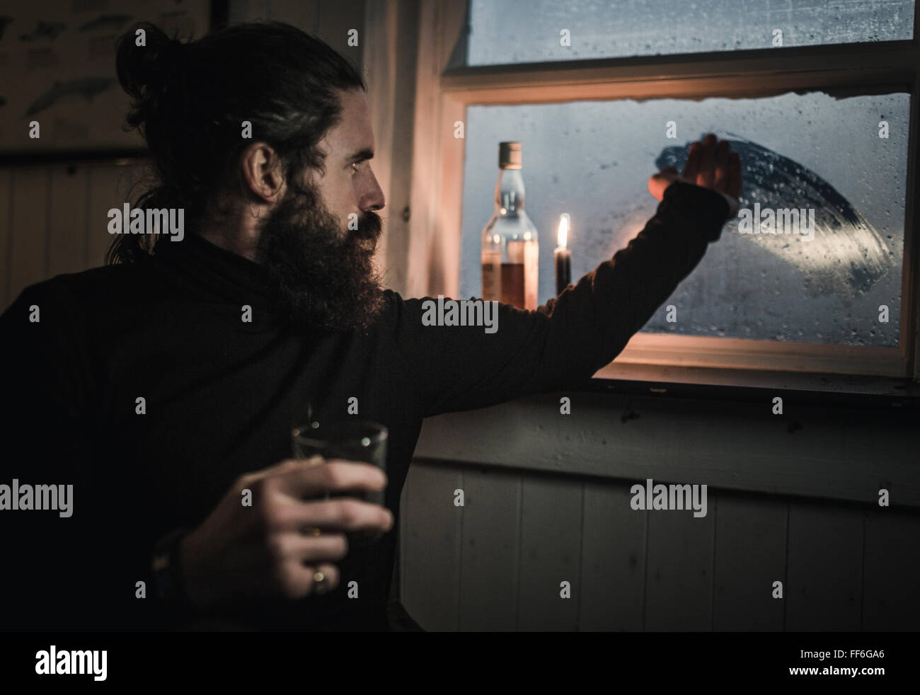 Ein Mann sitzt allein in einem Zimmer mit einer Flasche Whisky und ein Glas Fenster Kondenswasser abwischen Stockfoto