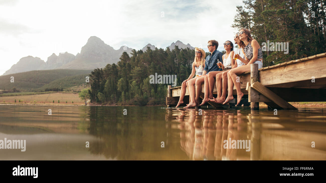 Porträt der glückliche junge Freunde sitzen auf Pier am See trinken Bier. Junge Männer und Frauen, die einen Tag am See zu genießen. Stockfoto