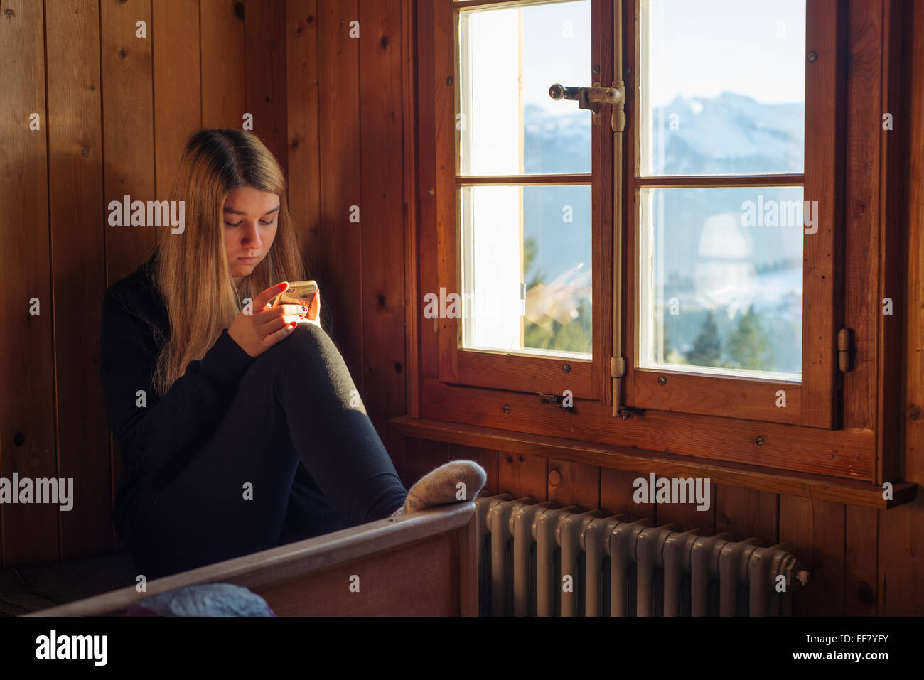 Junge Frau sitzt hinter dem Fenster im Inneren Berghütte mit Holzverkleidung Wand Smartphone betrachten Stockfoto